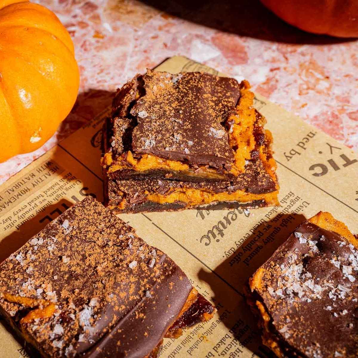 Three chocolate and pumpkin bars with sea salt are stacked on newspaper, surrounded by additional bars, a small pumpkin, and rustic date bark on a pink surface.