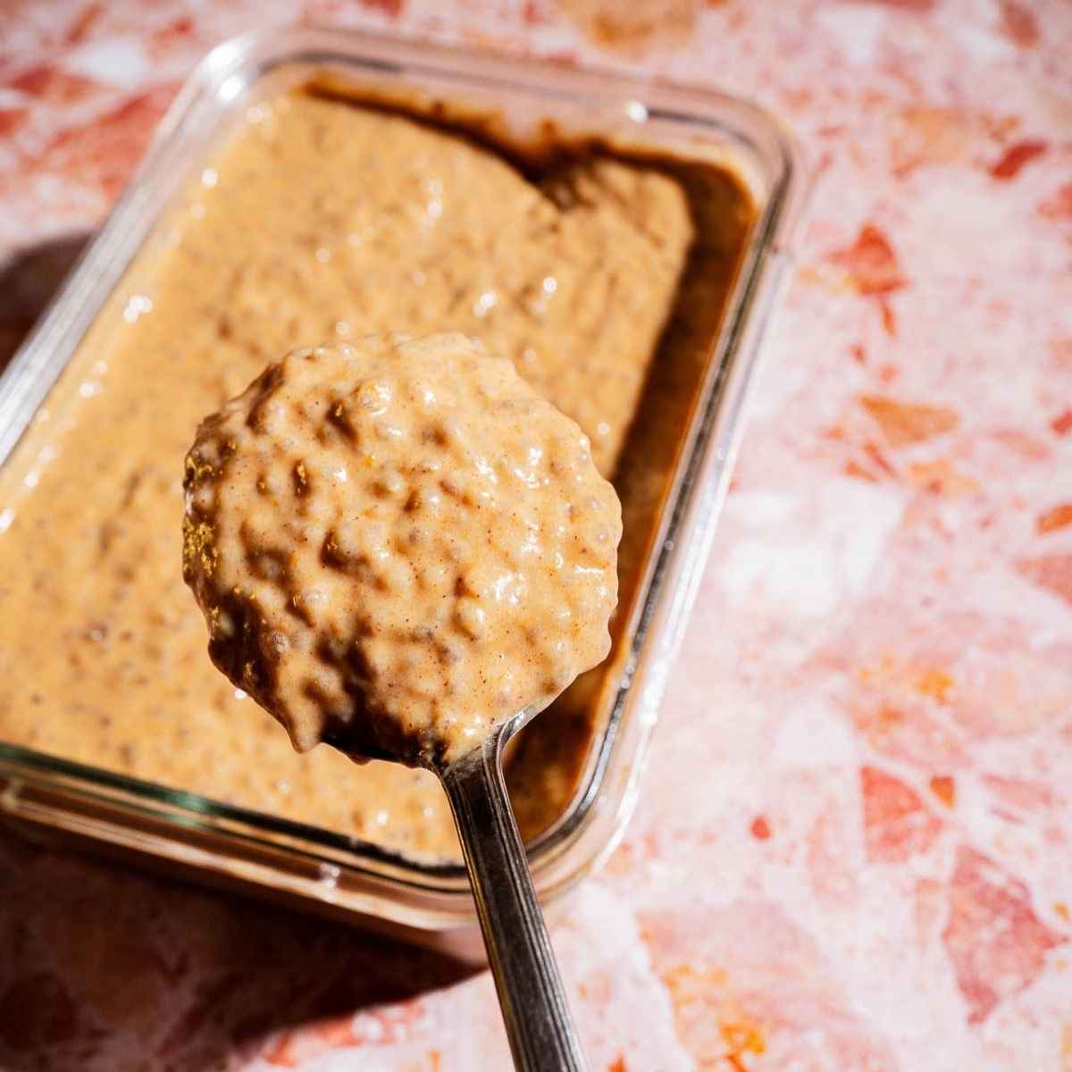 A spoon holds up a thick, tan pumpkin chia pudding mixture above a glass container filled with the same pudding, on a pink marbled surface.