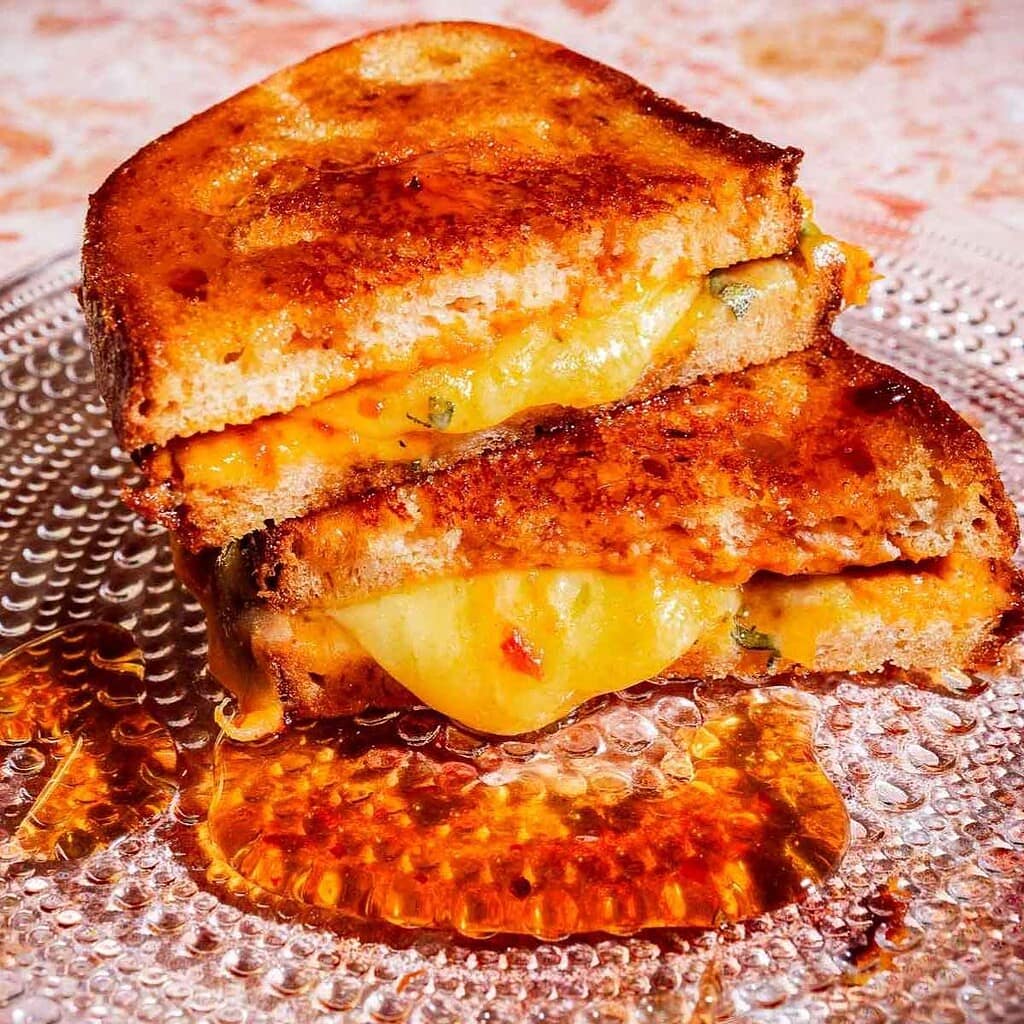 Two halves of a grilled cheese sandwich stacked on a glass plate with melted cheese and some oil or sauce underneath.