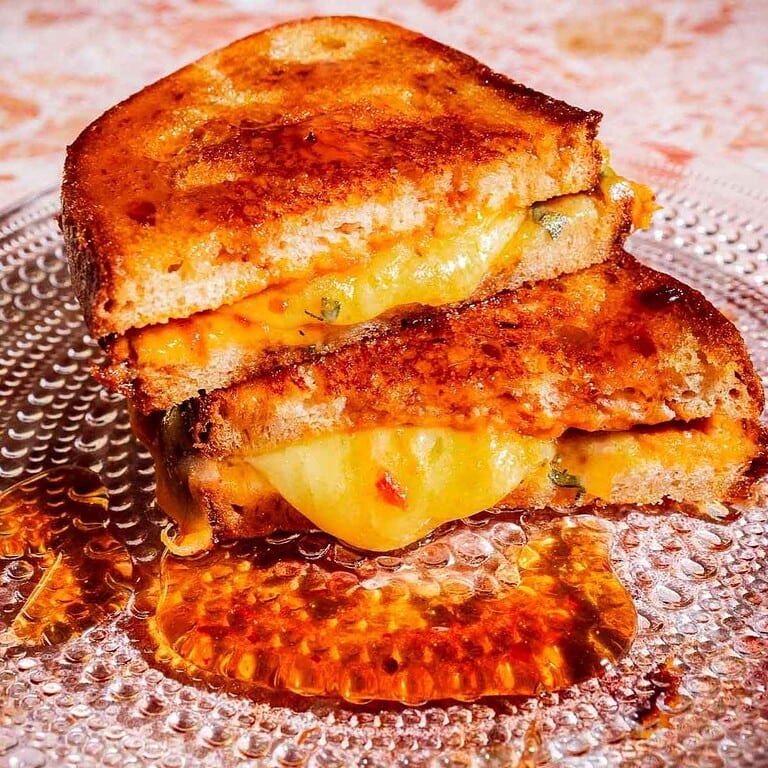 Two halves of a grilled cheese sandwich stacked on a glass plate with melted cheese and some oil or sauce underneath.
