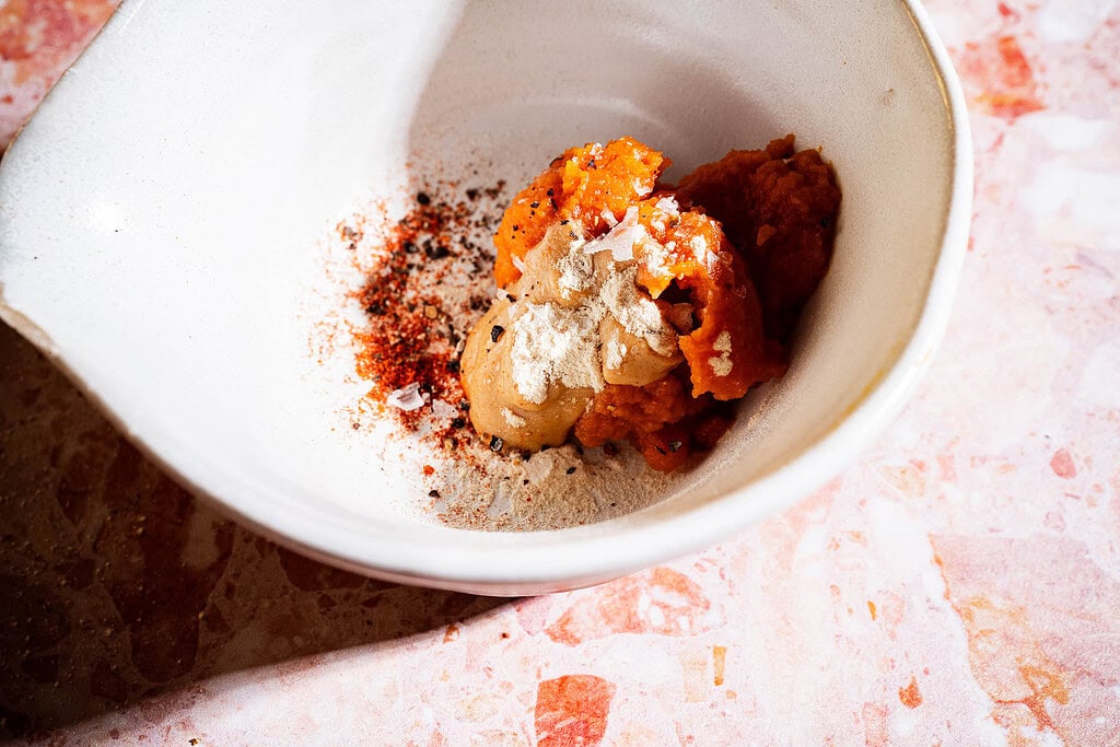 White bowl with scoops of orange puree, tan paste, chili powder, black pepper, and flaky salt on a pink marbled surface.