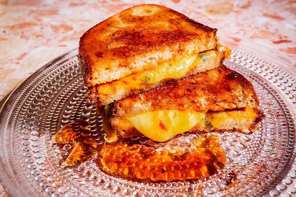 Two halves of a grilled cheese sandwich with melted cheese are stacked on a clear glass plate, with some cheese and grease pooling underneath.