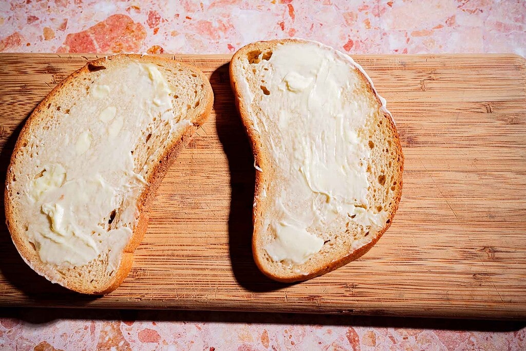 Two slices of white bread with butter spread on top are placed on a wooden cutting board with a pink speckled countertop underneath.