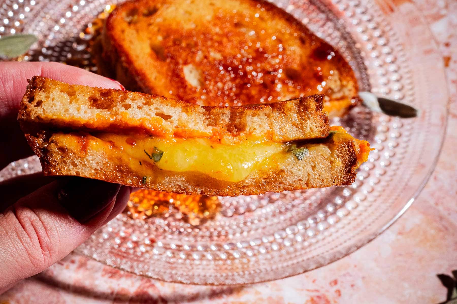 A hand holds a pumpkin grilled cheese sandwich with melted cheese and herbs, with another sandwich half on a clear glass plate in the background.