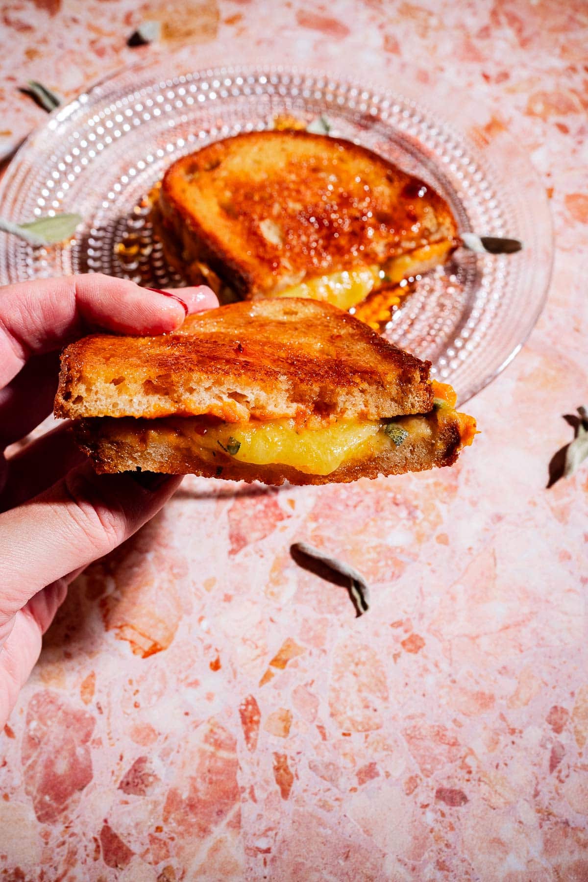 A hand holds a delicious pumpkin grilled cheese sandwich with melted cheese, while another sandwich half sits on a glass plate on a pink marble surface.