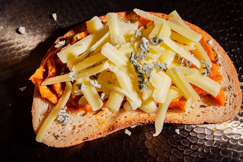 A slice of bread topped with orange spread, grated cheese, and sprinkled herbs sits on a textured dark surface.