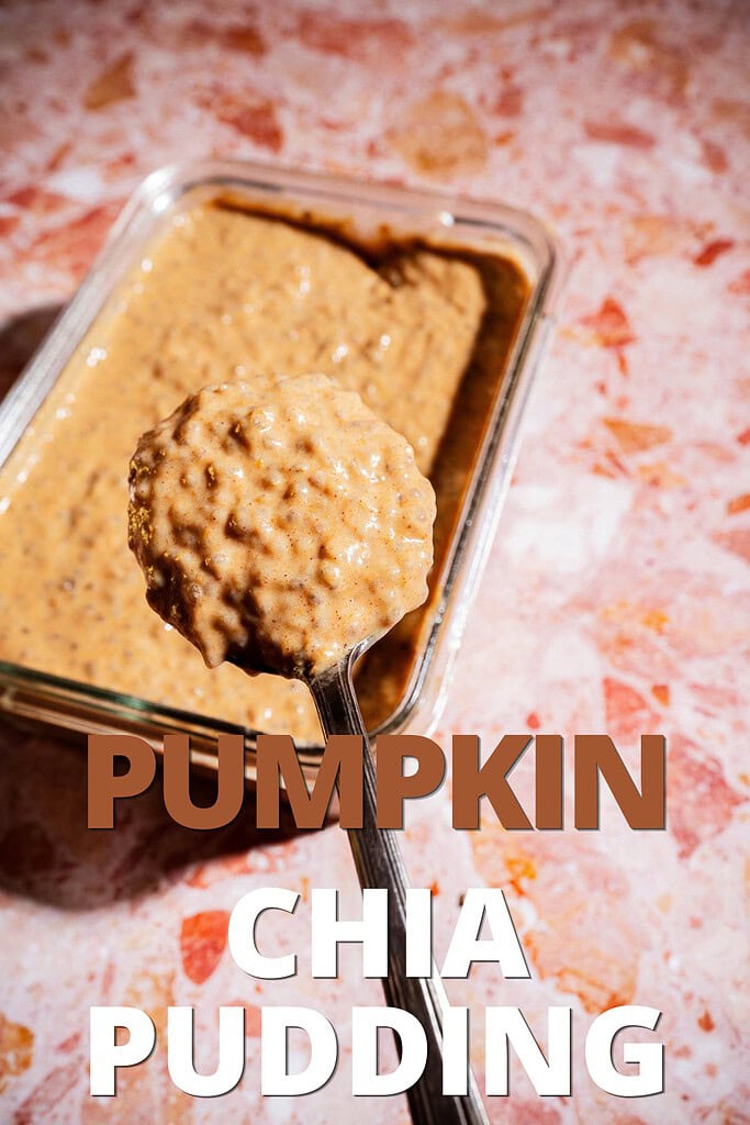 A spoonful of creamy pumpkin chia pudding is held above a glass container brimming with more of the autumn-inspired treat, all resting on a pink marbled surface. Text reads “PUMPKIN CHIA PUDDING.”.