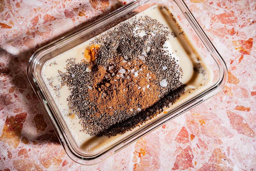 A glass dish contains milk, chia seeds, cocoa powder, and a few grains of salt on a pink marble surface, creating the perfect base for a rich pumpkin chia pudding.