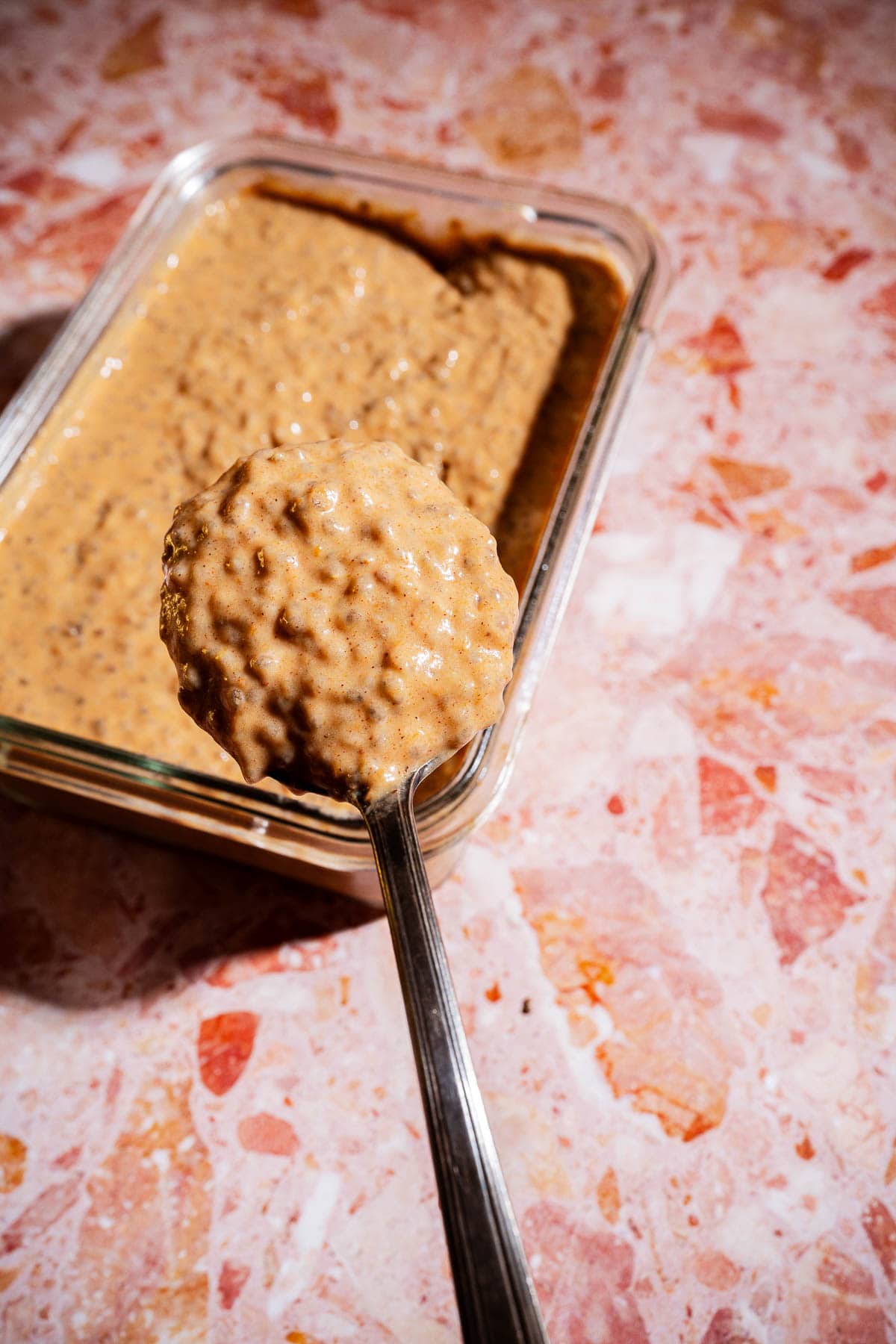 A spoon holding a scoop of thick, chunky pumpkin chia pudding mixture above a glass container, all on a pink and white marbled surface.