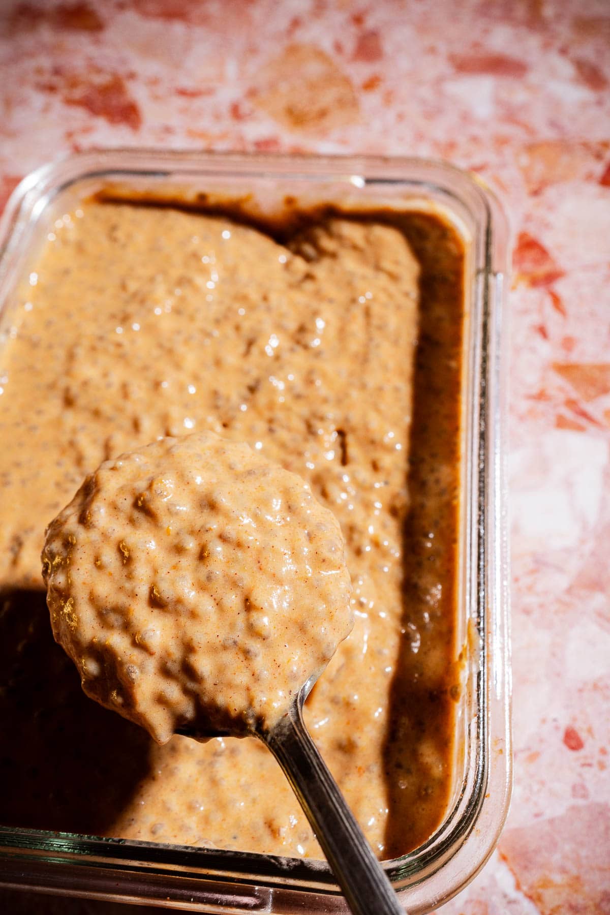 A spoon holding a portion of thick, brown pumpkin chia pudding hovers above a glass container filled with the same mixture on a marbled surface.