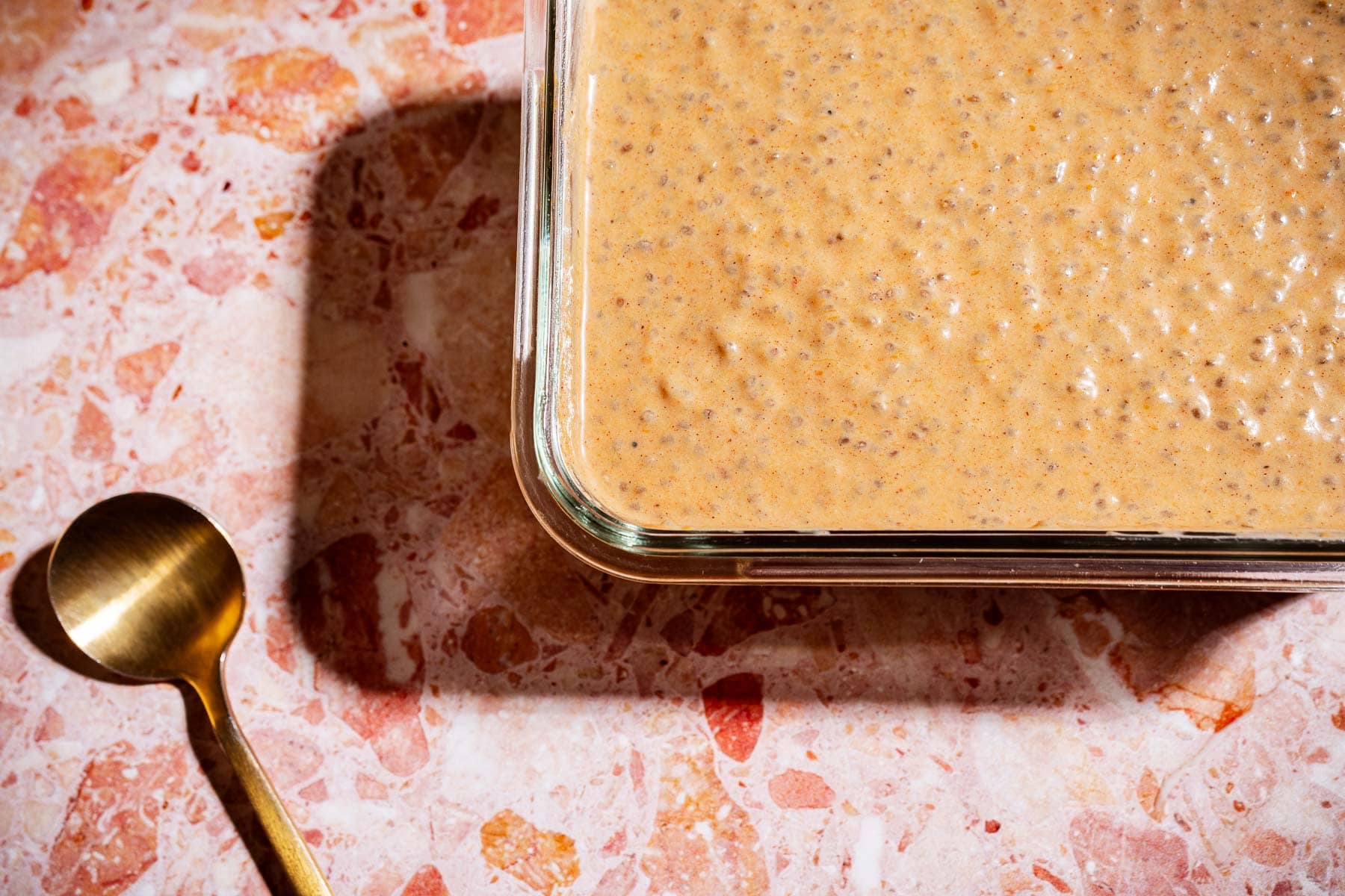 A glass dish filled with creamy, speckled pumpkin chia pudding sits on a pink marbled surface next to a gold spoon.