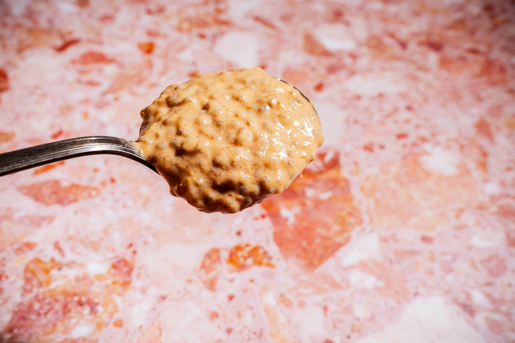 A metal spoon holds a heaping scoop of chunky, light brown peanut butter against a pink and white marbled background, ready to swirl into your favorite pumpkin chia pudding.