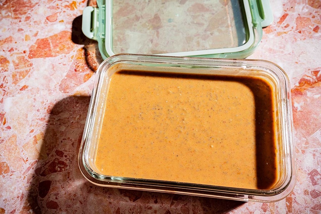 A glass container filled with a thick, brownish-orange pumpkin chia pudding sits on a pink marbled surface with its green-lidded cover open.