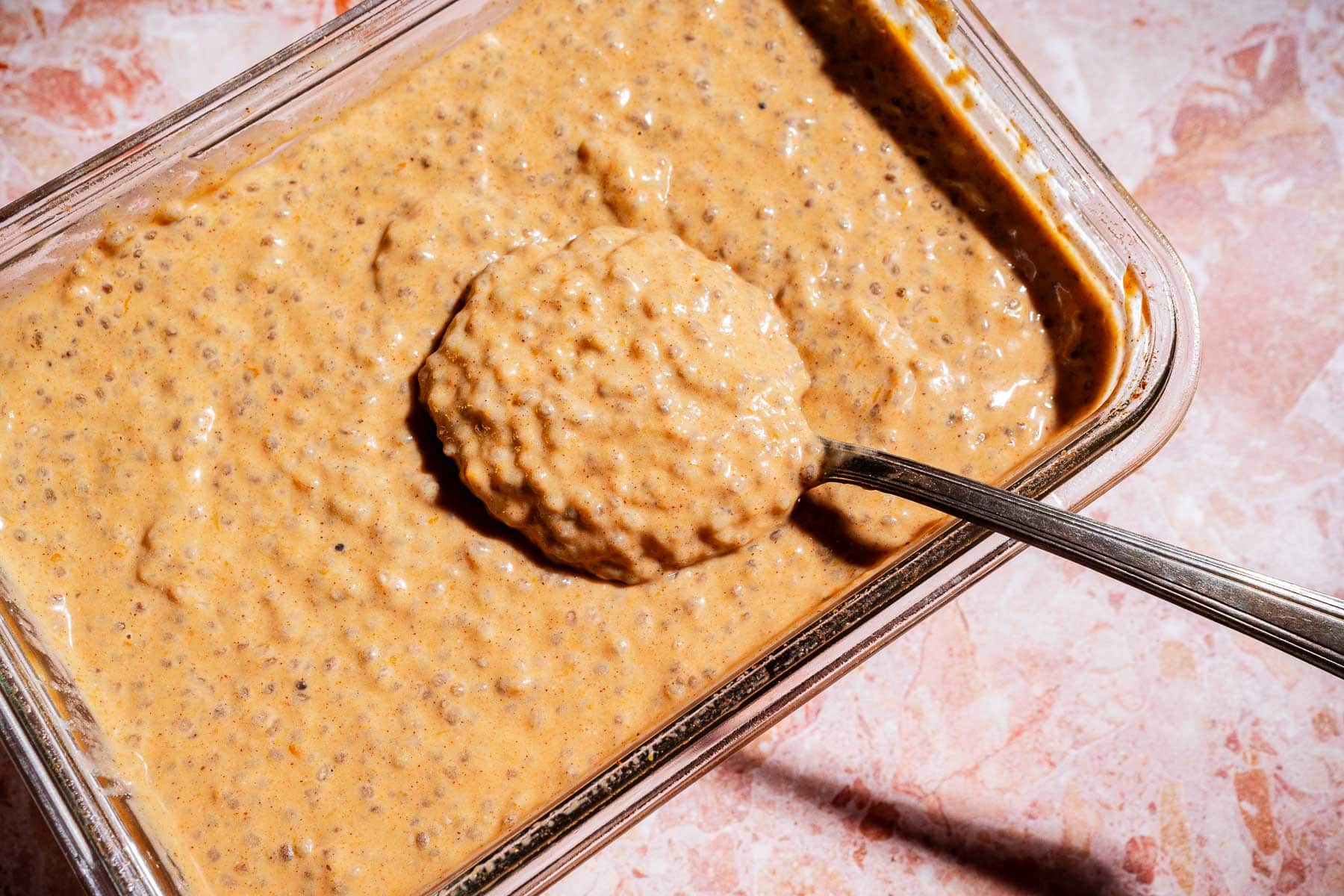 A metal spoon scoops thick, orange-brown pumpkin chia pudding from a rectangular glass container on a marble surface.