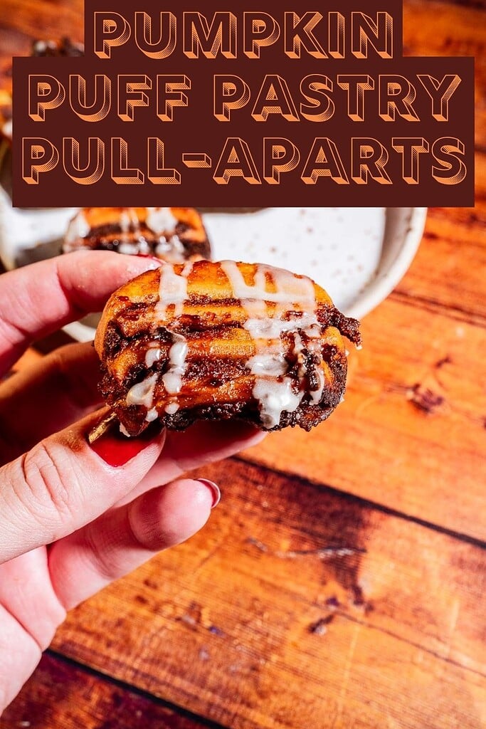 A hand holds a small pumpkin puff pastry pull-apart topped with icing, with more pastries on a plate in the background on a wooden surface.