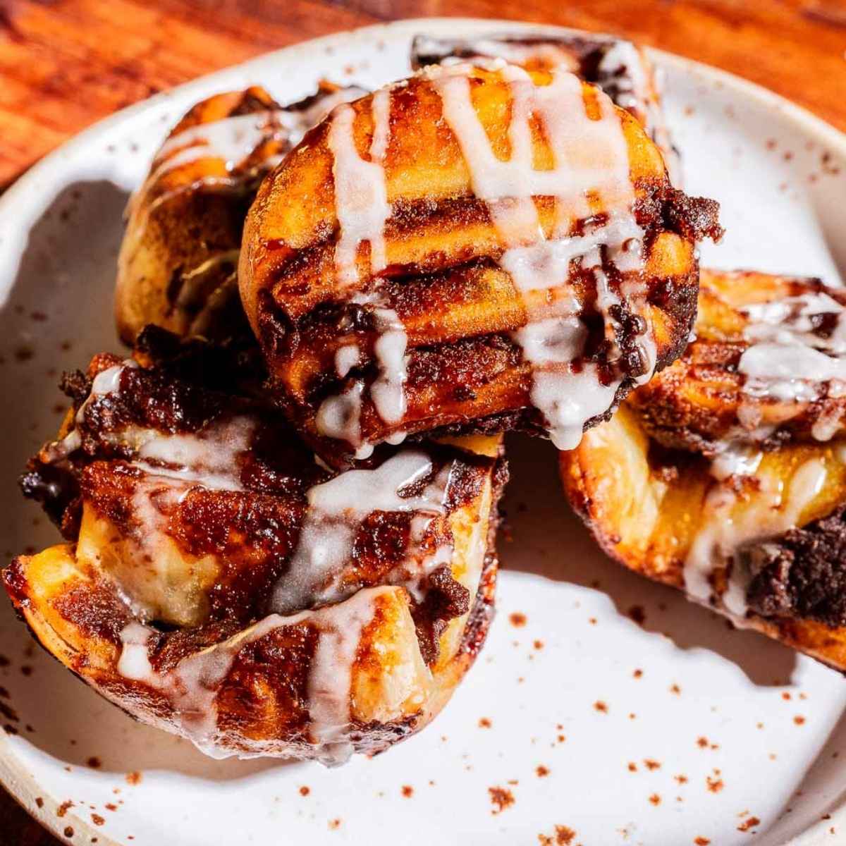 Three pumpkin pastries with icing drizzle are stacked on a white plate with brown speckles, set on a wooden surface.