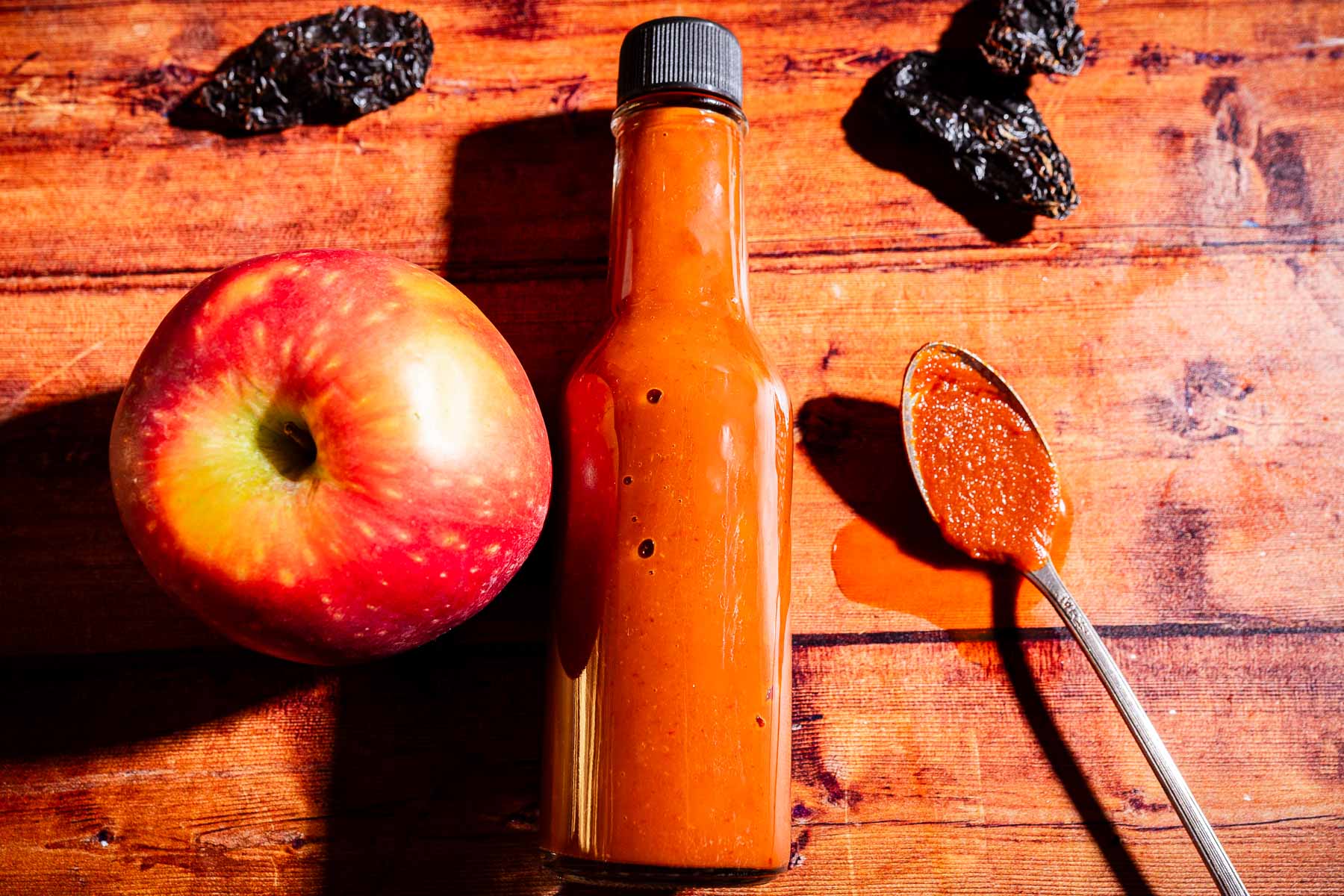 A bottle of apple hot sauce, a fresh apple, dried peppers, and a spoon with sauce are arranged on a wooden surface.
