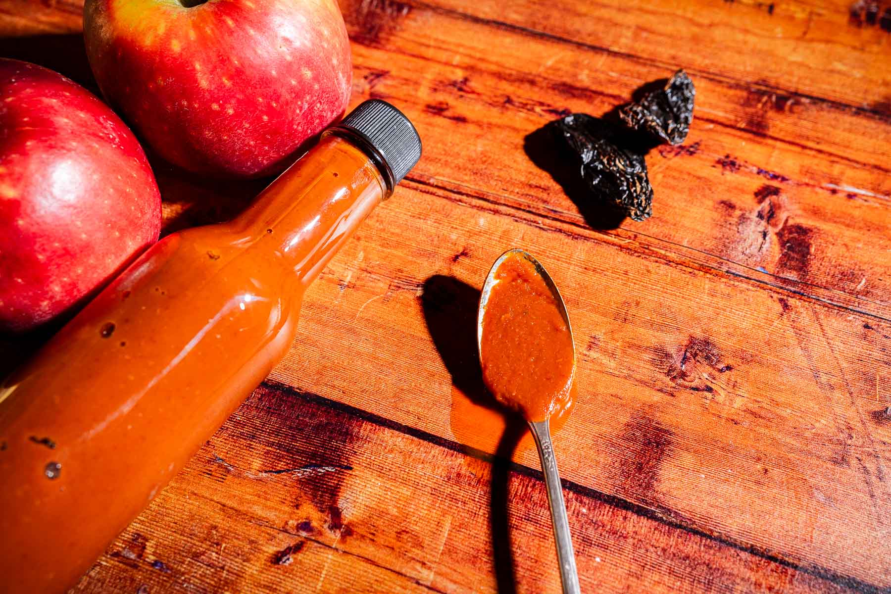 A bottle of reddish apple hot sauce, a spoon with sauce, two apples, and dried chili peppers on a wooden surface.