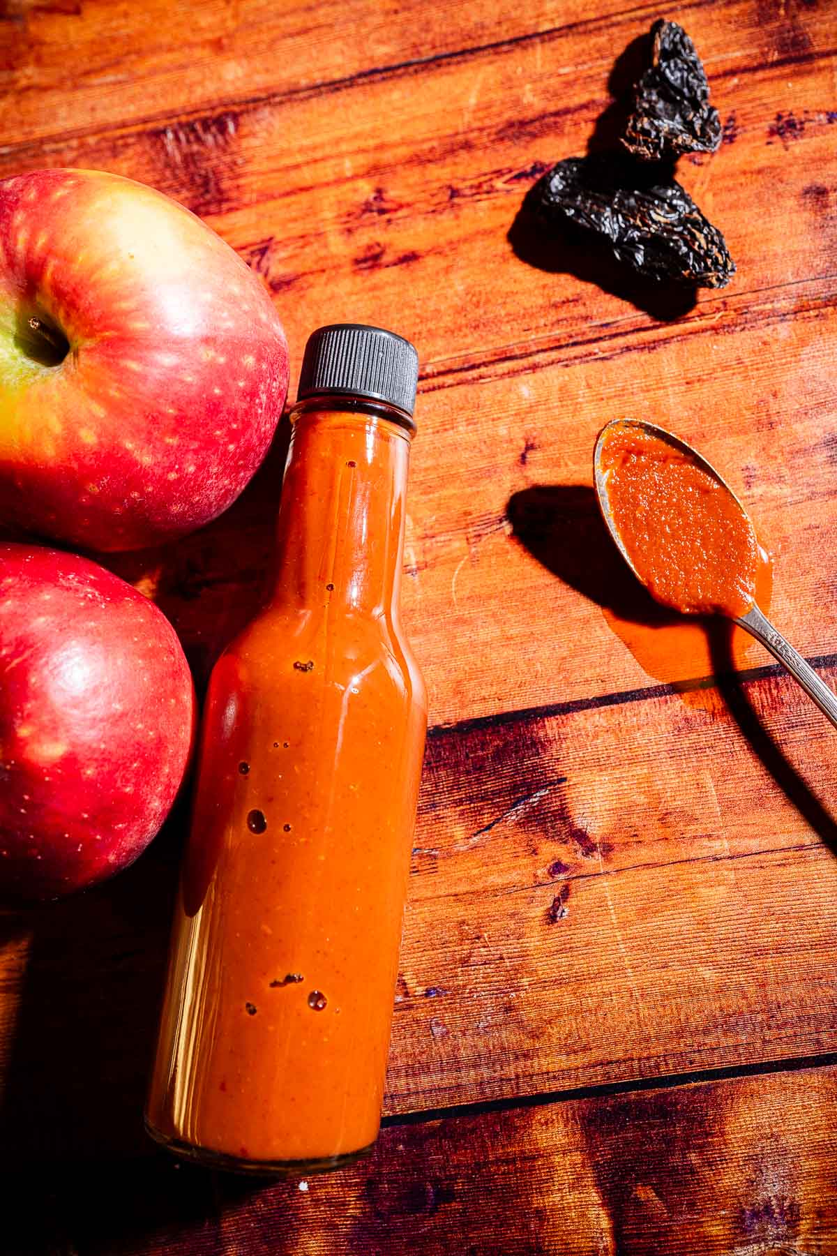 A bottle of reddish-brown apple hot sauce sits on a wooden surface, accompanied by two apples, dried peppers, and a spoonful of sauce beside it.