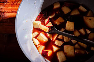 White bowl with diced apples in a reddish apple hot sauce, possibly a marinade or tangy condiment, with a metal spoon resting inside. The bowl is partially lit, highlighting the ingredients.