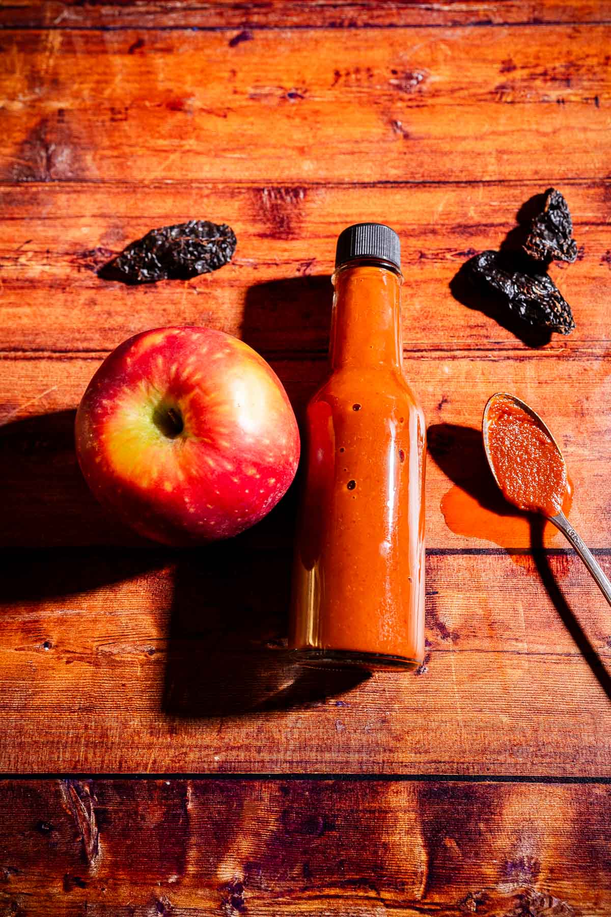 A bottle of reddish apple hot sauce sits on a wooden surface next to a red apple, a spoon with sauce, and three dried peppers.