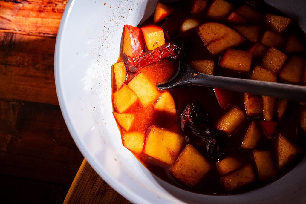 Bowl with cubed fruit and dried chilies in a red apple hot sauce, stirred with a black spoon, on a wooden surface.