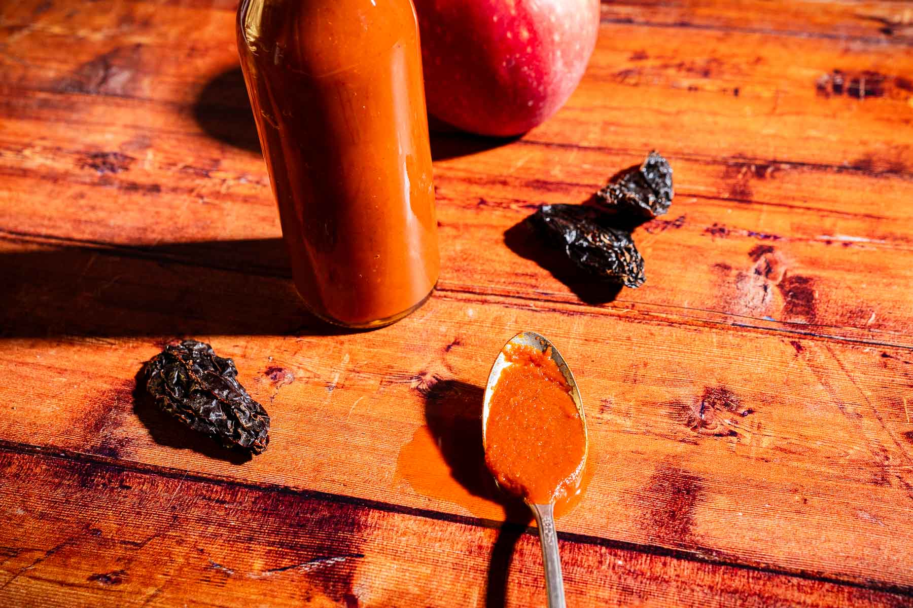 A bottle of reddish apple hot sauce, a spoon with sauce, dried peppers, and an apple rest on a wooden surface under bright light.