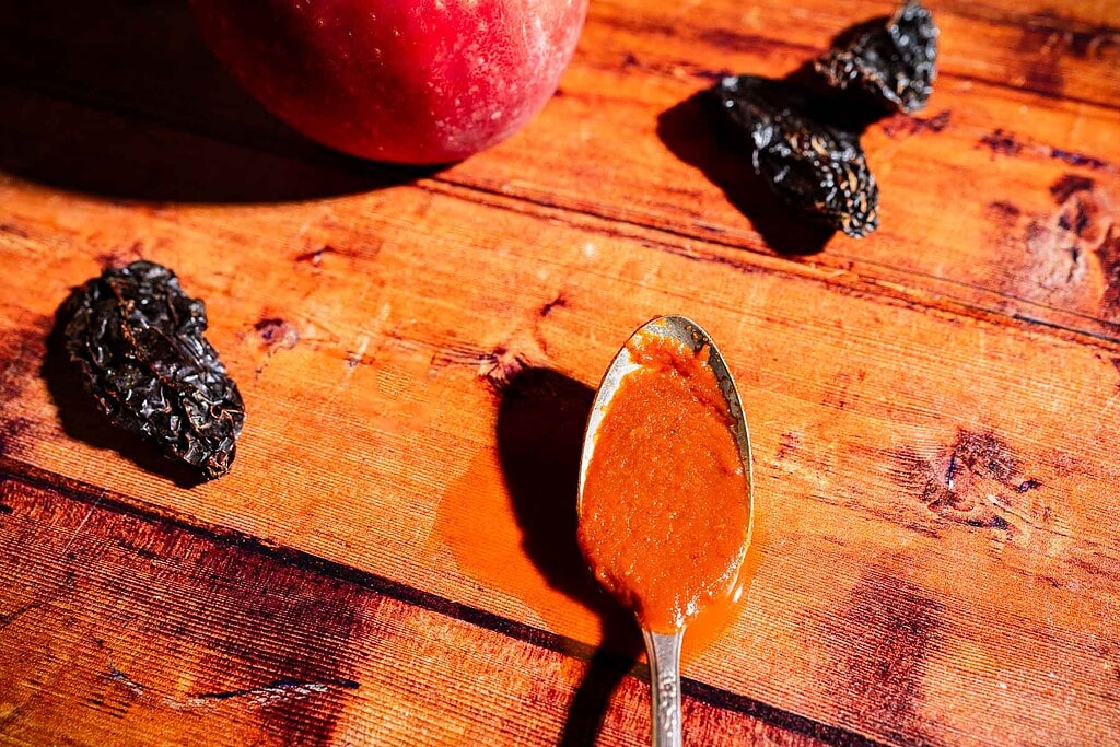 A spoon with reddish-brown apple hot sauce on a wooden table, next to dried chilies and a whole peach.