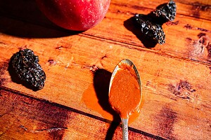 A spoon with reddish-brown apple hot sauce on a wooden table, next to dried chilies and a whole peach.