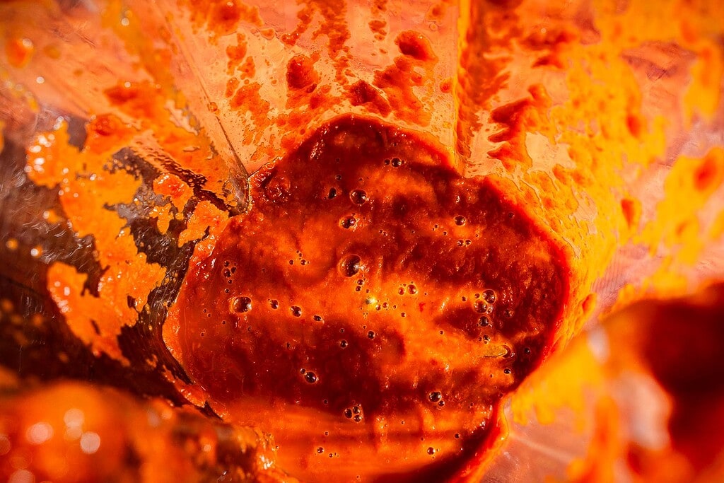 Close-up of thick, red-orange apple hot sauce with bubbles and streaks on the sides of a blender container.