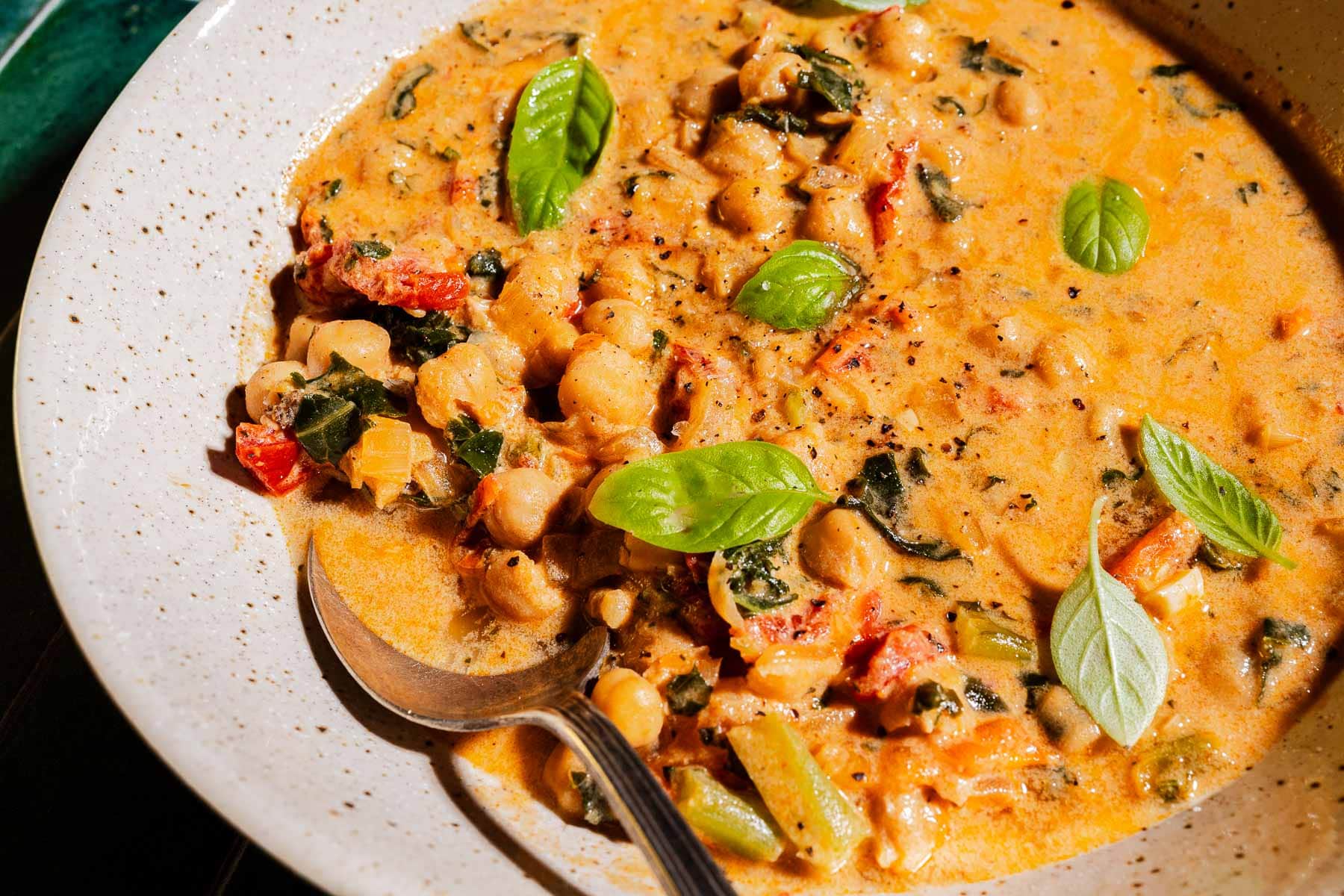 A bowl of creamy chickpea soup with spinach, tomatoes, and fresh basil leaves, served with a spoon.