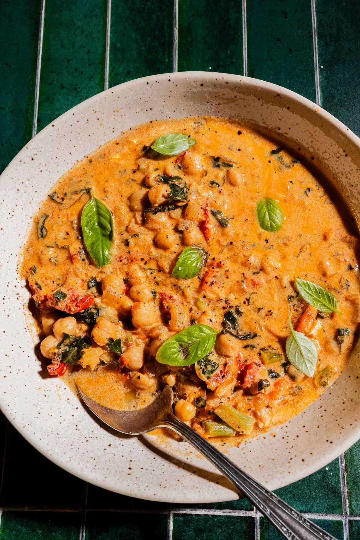 A bowl of creamy chickpea soup with greens and tomato, garnished with fresh basil leaves, sits on a green tiled surface with a spoon.