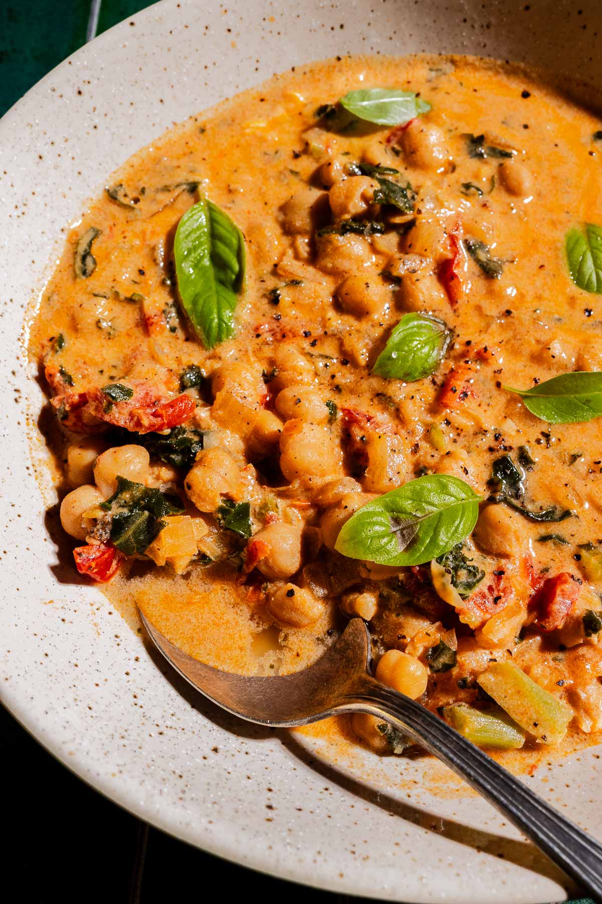 A bowl of creamy chickpea soup with spinach, sun-dried tomatoes, and fresh basil leaves, served with a spoon.