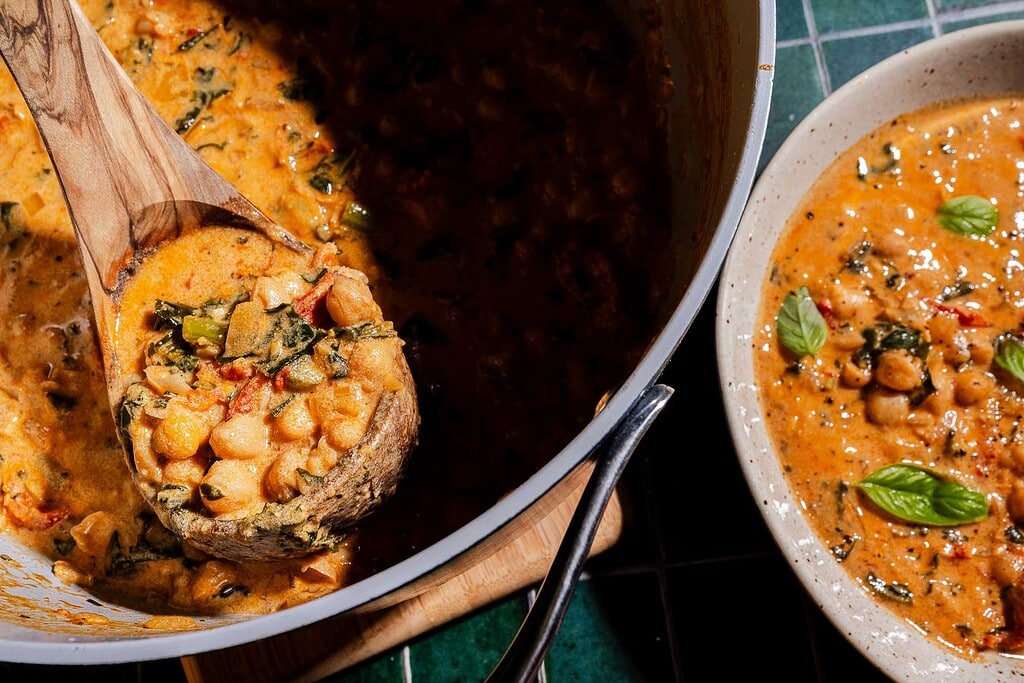 A pot of creamy chickpea soup with vegetables is being stirred with a wooden spoon; a bowl of the soup sits beside the pot, garnished with basil leaves.