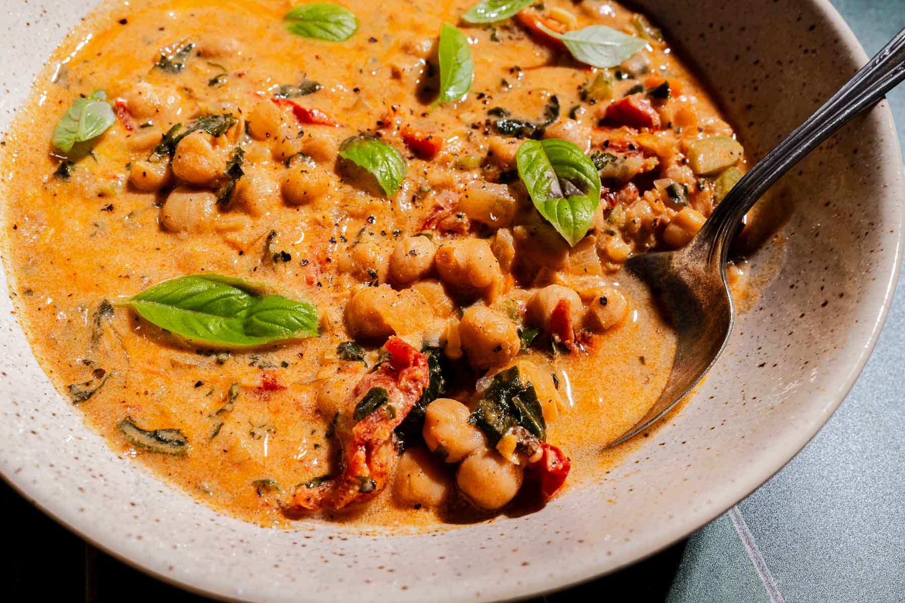 A bowl of creamy chickpea soup with visible chickpeas, sun-dried tomatoes, fresh basil leaves, and a spoon resting inside the bowl.