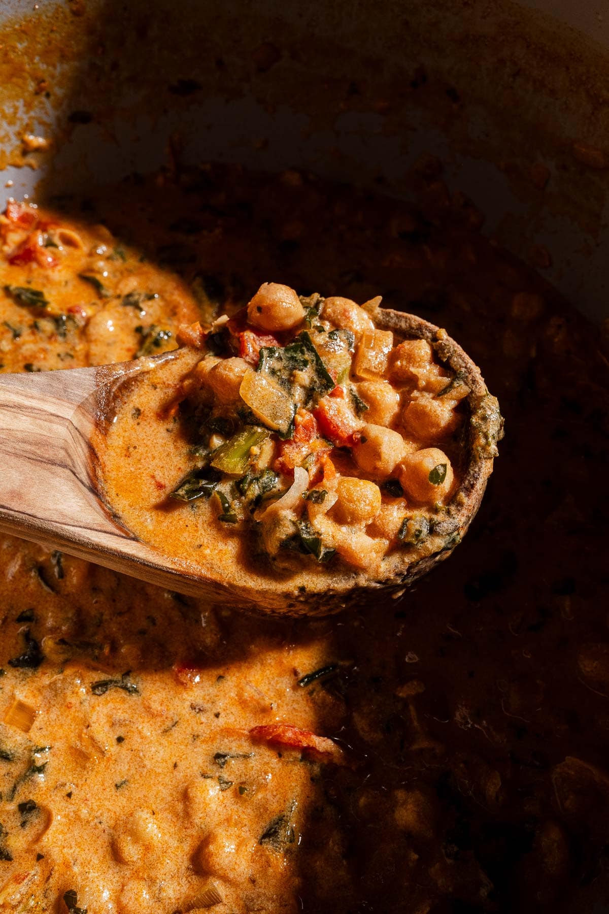 A wooden spoon holds a portion of creamy chickpea soup and vegetable stew above a pot filled with the same dish.