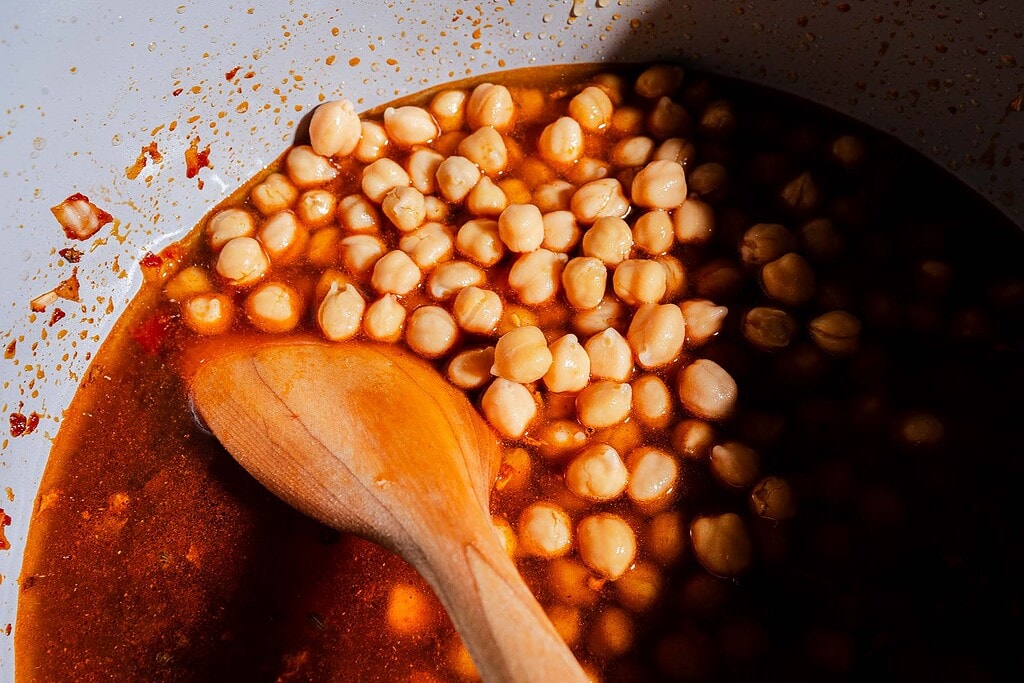 Wooden spoon stirring chickpeas in a red, oily liquid inside a white pot, with some shadow covering part of the hearty chickpea soup.