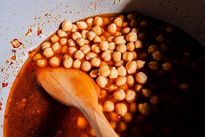 Wooden spoon stirring chickpeas in a red, oily liquid inside a white pot, with some shadow covering part of the hearty chickpea soup.