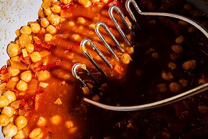 A potato masher pressing chickpeas in a tomato-based chickpea soup with onions and diced vegetables, under bright lighting.