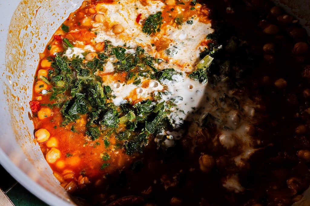 A close-up of a pot containing a hearty chickpea soup with tomato sauce, greens, and cream, partially in shadow.