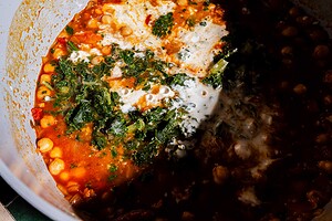 A close-up of a pot containing a hearty chickpea soup with tomato sauce, greens, and cream, partially in shadow.
