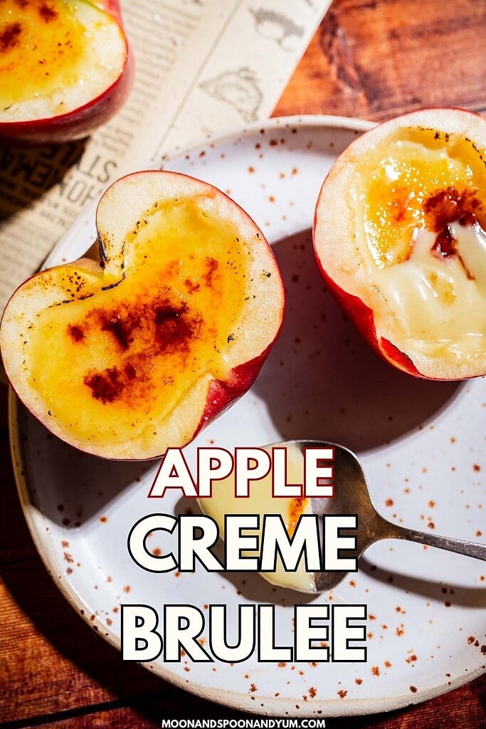 Halved apples filled with a luscious apple creme brulee topping, lightly caramelized, are served on a white speckled plate with a spoon atop a wooden table. Text overlay reads “Apple Creme Brulee.”.