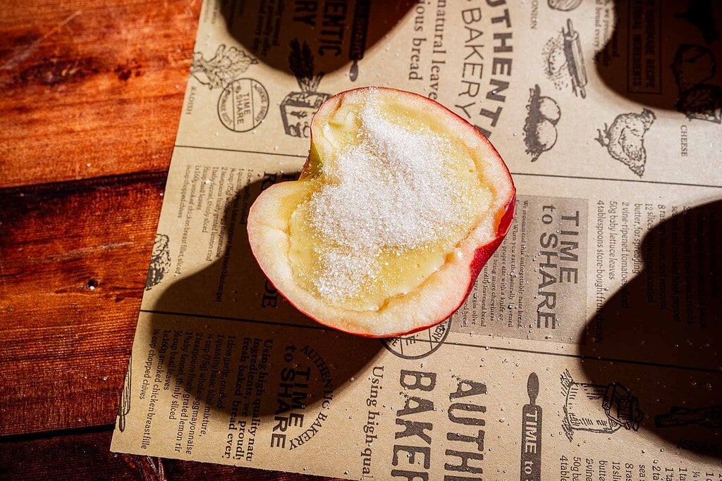 Half of an apple with its core removed, filled with sugar for a delightful apple creme brulee twist, placed on decorative parchment paper on a wooden surface.
