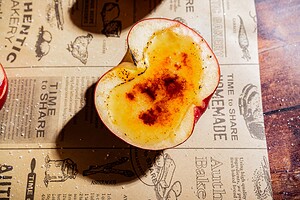A halved apple creme brulee with a caramelized, seasoned surface sits on brown parchment paper adorned with bakery-themed illustrations and text.