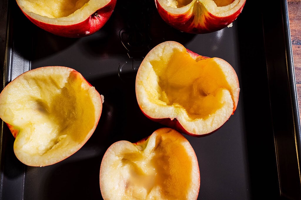 Hollowed-out apple halves sit on a black baking tray, their empty centers ready to be transformed into a delightful apple creme brulee, prepared for filling or baking.