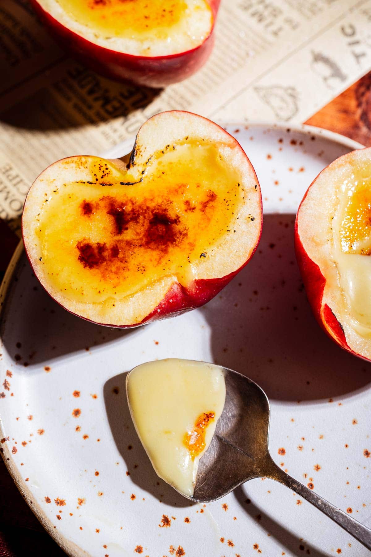A halved apple creme brulee filled with custard and caramelized sugar sits on a plate with a spoonful of custard beside it, on top of a newspaper.