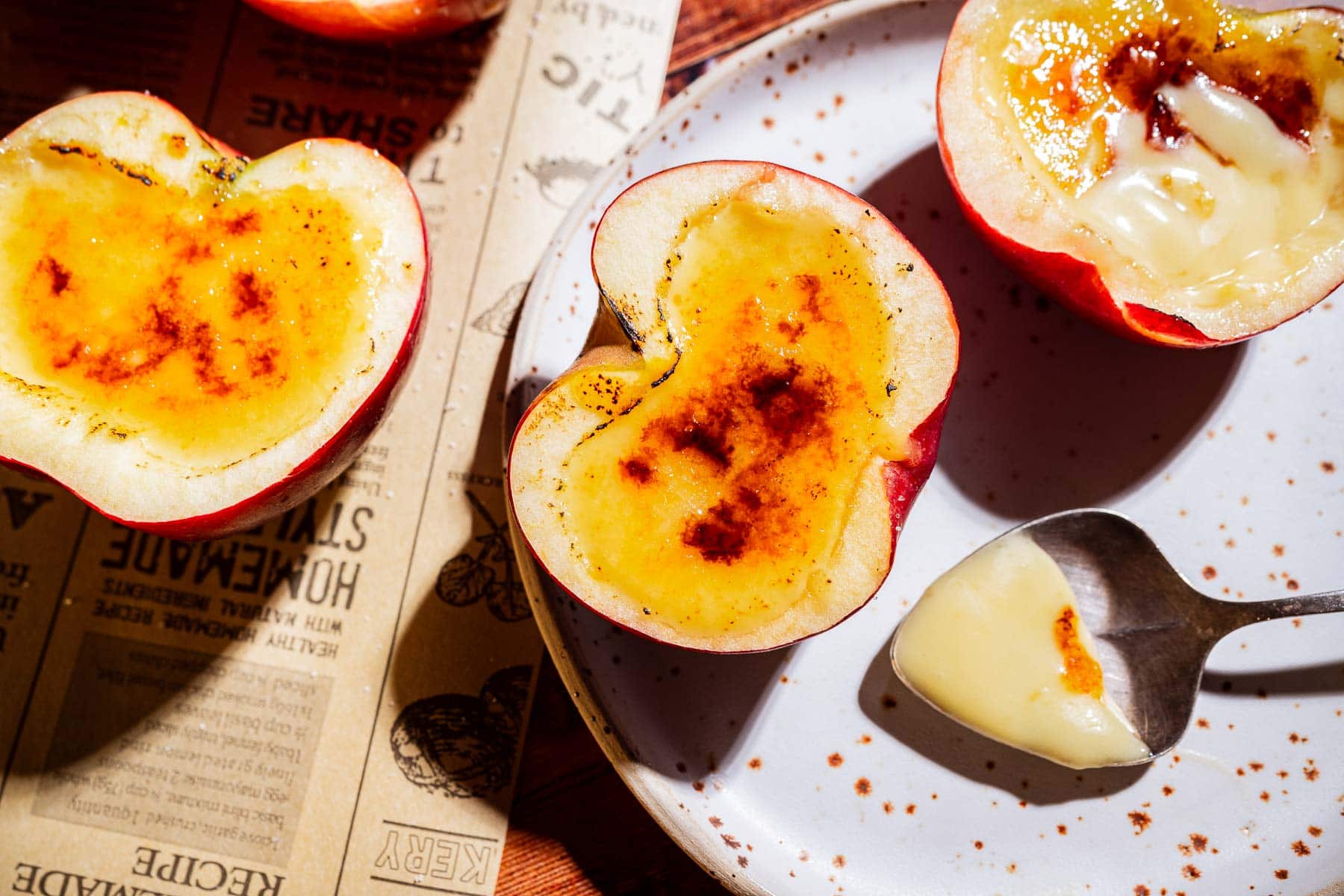 Halved plums with brûléed tops are plated with a spoonful of apple creme brulee custard on a white speckled dish; a newspaper rests beneath the fruit.