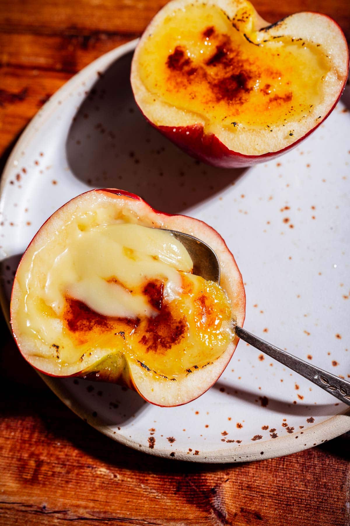 Two halved apples with a creamy, caramelized apple creme brulee filling sit on a speckled white plate; a spoon rests in one half.