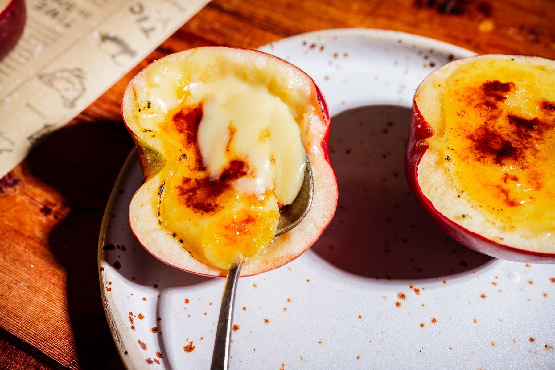 A halved apple filled with creamy custard, lightly torched on top to create a delicate caramelized crust, sits on a white speckled plate with a spoon scooping some filling—an inventive twist on classic apple creme brulee.