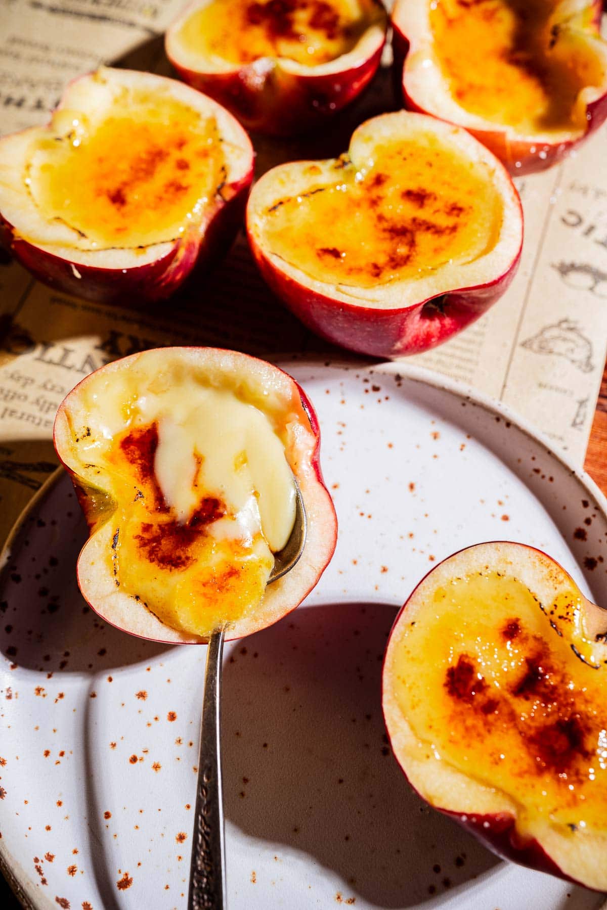 Halved apples filled with creamy apple creme brulee and caramelized sugar, arranged on a speckled white plate atop a newspaper-patterned surface, with a spoon scooping custard from one half.